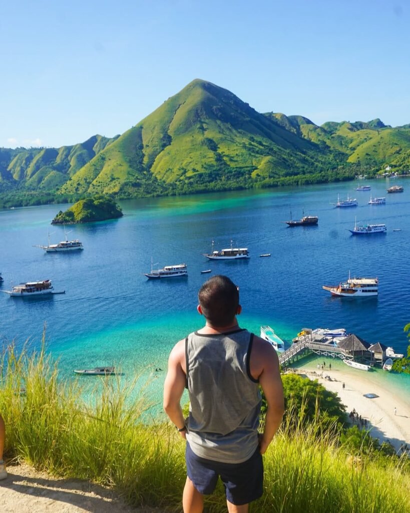 Turis menikmati pemandangan indah dari puncak Pulau Kelor dalam paket liburan Labuan Bajo, dengan laut biru dan gugusan pulau di latar belakang